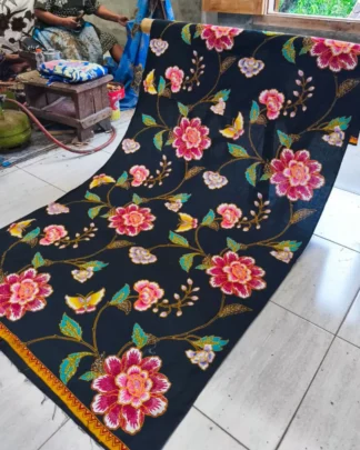 Kain batik sarung wanita motif bunga-bunga cerah di atas dasar kain hitam, dengan seorang pembatik di latar.