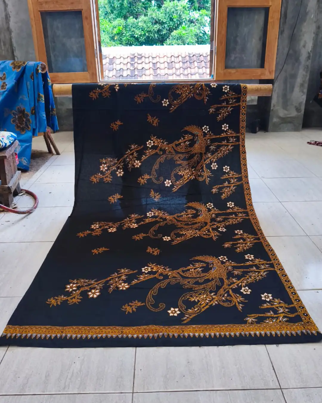 Kain batik sarung wanita motif Burung Paksi Sogan yang anggun di atas dasar kain hitam klasik, dengan seorang pembatik di latar belakang.