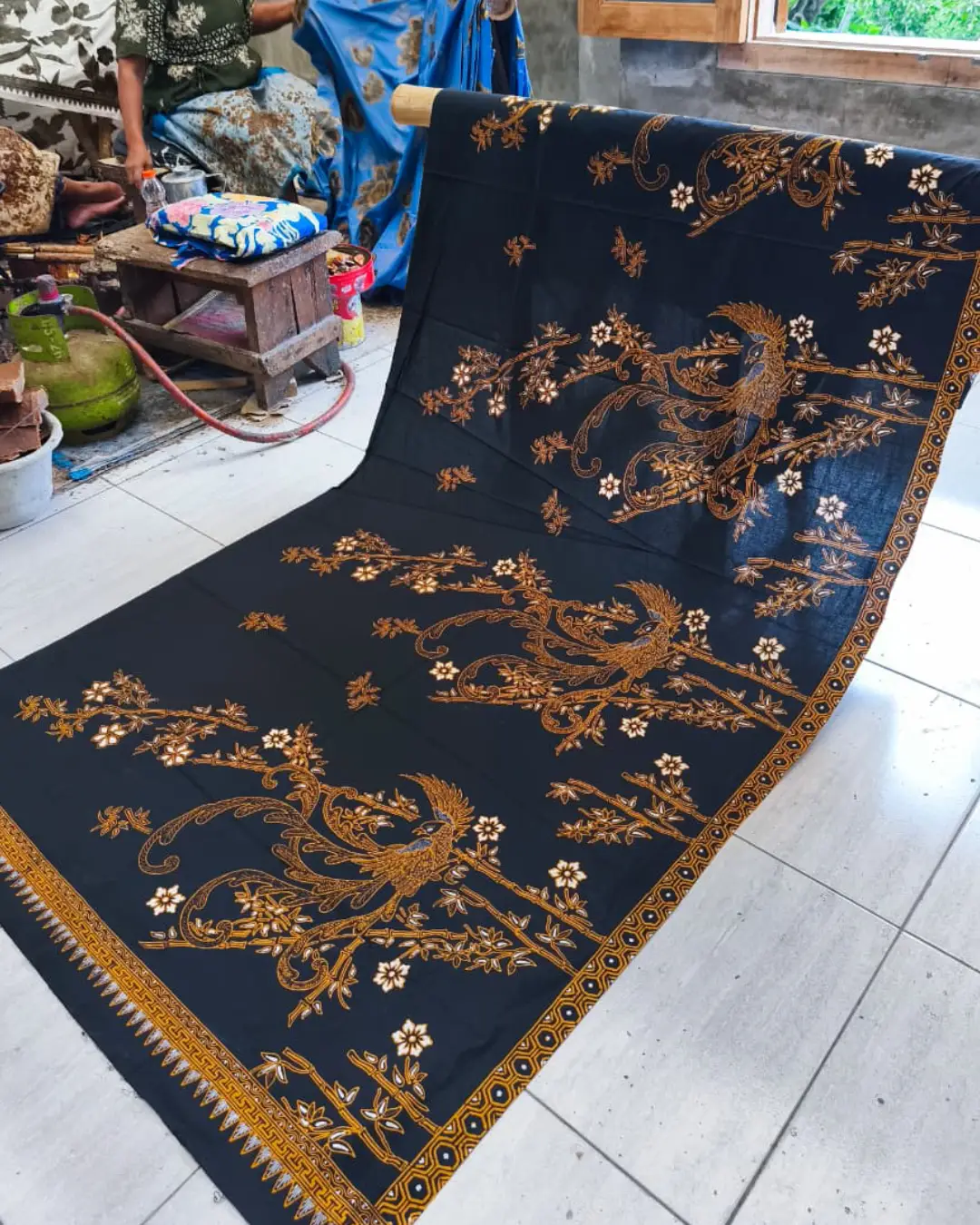 Kain batik sarung wanita motif Burung Paksi Sogan yang anggun di atas dasar kain hitam klasik, dengan seorang pembatik di latar belakang.